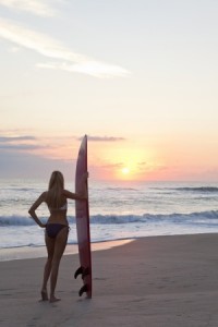 Rear view of a beautiful young woman surfer girl in bikini with surfbord at a beach at sunset or sunrise