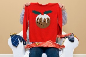 A man opening Christmas presents to discover he got a Christmas themed jumper to go along with the usual socks and tie