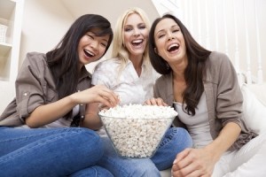 Three beautiful young women friends at home eating popcorn watching a movie together and laughing