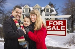Warmly Dressed Young Mixed Race Family in Front of Home For Sale Real Estate Sign and House with Snow On The Ground