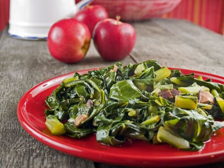 Collard greens and bacon on a red plate with apples and a water pitcher in the background