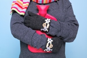 Photo of a woman holding a red hot water bottle to her chest whilst wearing hand kniited woolen gloves trying to keep warm, good image for winter illness or warmth related themes.