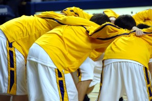 High school varsity basketball team huddles together before game start. Uniforms are yellow, purple and white.