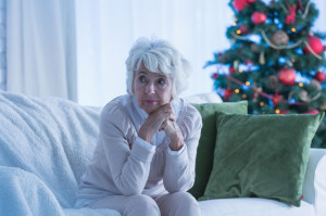65562202 - senior woman sitting alone on sofa, christmas tree in background