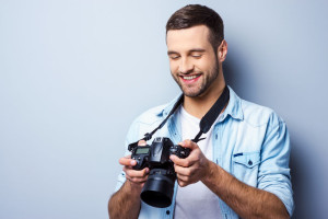 Great shot! Handsome young man holding digital camera and looking at it with smile while standing against grey background