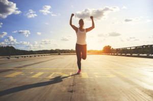 Winner as style of life. Horizontal shot of young beautiful woman in sports clothing keeping arms raised and smiling while passing finish line during jogging. Evening sunlight on background.