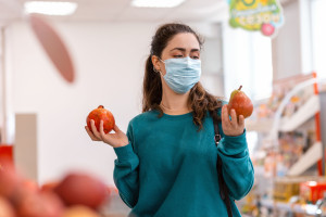 Shopping. Portrait of a young Caucasian woman with a medical mask on her face, holding a pear and a pomegranate, choosing between them. The concept of buying fruit and protecting against coronovirus