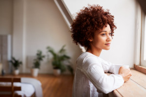 Young Woman Relaxing In Loft Apartment Looking Out Of Window With Hot Drink