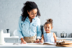 smiling african american mother and daughter looking at cookbook together in kitchen