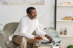 happy african american man sitting in armchair and listening music on record player