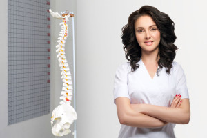Young pretty female doctor in uniform keeping arms crossed and looking at camera. Artificial human cervical spine model in medical office. Orthopedic practice