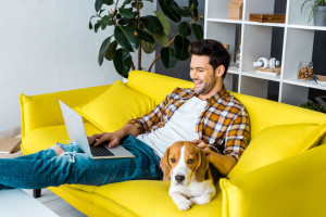 handsome laughing man using laptop on sofa with beagle dog