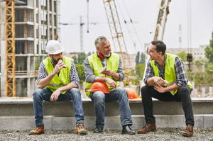 Time for a break. Group of builders in working uniform are eating sandwiches and talking while sitting on stone surface against construction site. Building concept. Lunch concept