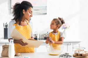 african american mother holding cookbook and daughter preparing dough in kitchen, looking at each other