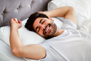 Happy wellslept arab man awakening and stretching hands lying in modern comfy bed at home