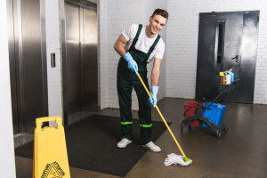 handsome young janitor mopping floor and smiling at camera