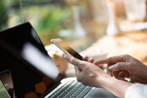 Woman hand holding white mobile phone on a table with a laptop i