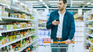 At the Supermarket: Handsome Man Uses Smartphone and Looks at Nutritional Value of the Canned Goods. He's Standing with Shopping Cart in Canned Goods Section.