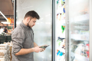 Bearded man with frozen food in his hands stands at the refrigerator in the department of frozen food. Buyer selects and purchases frozen food in the grocery store. Shopping supermarket.