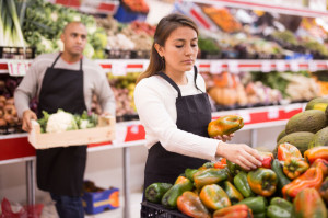 Latin saleswoman in black apron and her assistant with box of vegetables