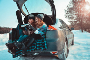Happy friends in winter forest. Two girls sit in trunk of car drinking coffee