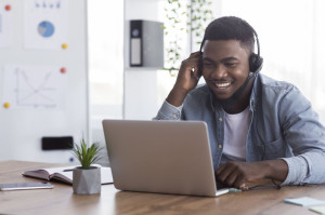 Black worker wearing headphones, watching webinar on laptop in office