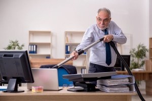 Old businessman employee holding vacuum cleaner at workplace