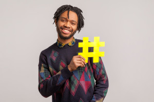 African man with dreadlocks and toothy smile on his face holding hashtag symbol, recommending popular topics, internet trends