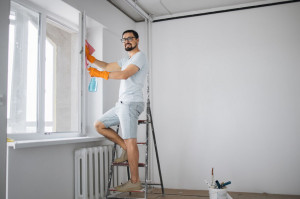 Attractive bearded man sitting on stepladder holding sponge for washing windows and detergents