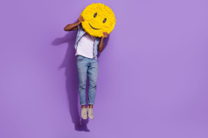 Full length photo of positive cheerful man wear jeans jumping empty space holding smile pinata isolated violet color background