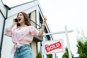 low angle view of cheerful woman gesturing while talking on smartphone near sold lettering on board