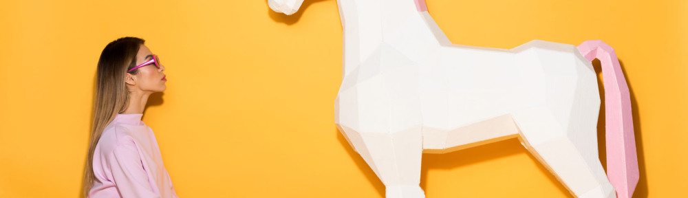 side view of asian female model in sunglasses sitting on floor and decorative unicorn on yellow background
