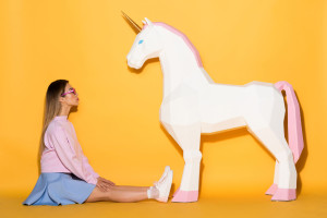 side view of asian female model in sunglasses sitting on floor and decorative unicorn on yellow background
