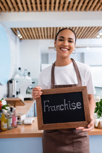 Young african american saleswoman holding chalkboard with franchise lettering in sweet shop