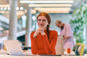 In a modern startup office, a professional businesswoman with orange hair sitting at her laptop, epitomizing innovation and productivity in her contemporary workspace.