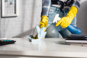 Cropped view of woman in rubber gloves cleaning table with rag and detergent near calculator on blurred foreground