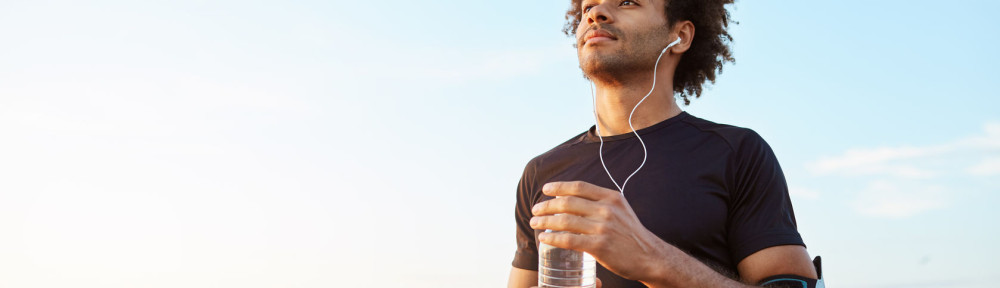 Man athlete drinking water out of plastic bottle after hard running workout. Dark-skinned male sportsman looking at the sky while running, enjoying view