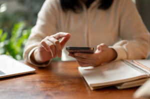 Cropped image of an Asian woman in casual clothes using her smartphone at a table indoors.