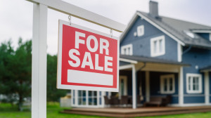 Close Up of a Red Real Estate Property for Sale Flopping in the Wind. Background with a Big Blue Stylish Countryside House. Housing Market Con