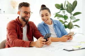 Gadgets concept. Two colleagues, man and woman sitting at a desk at office, using smartphones during working time, messaging online with clients or friends, reading news, searching information, smile